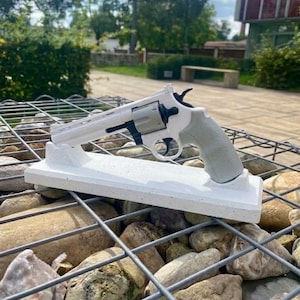 May include: A white and grey revolver pistol on a white rectangular stand. The gun has a grey grip and a black trigger. The stand rests on a bed of rocks within a wire mesh structure.