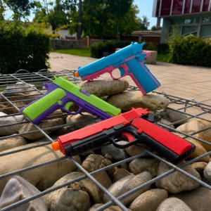 May include: Three toy pistols in bright colour combinations: one blue and pink, one lime green and purple, and one red and black. Each toy features a textured grip, a trigger, and a small orange tip at the end of the barrel, resting on a metal grid.