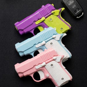 Peut inclure: Trois pistolets jouets aux couleurs pastel : violet, bleu clair et rose. Chacun a une poignée blanche avec un motif texturé et des accents contrastés de vert ou de blanc. Une télécommande de voiture est en arrière-plan.