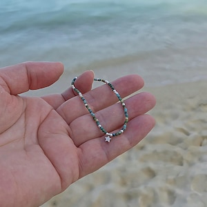 Puede incluir: Una delicada pulsera de cuentas con pequeñas cuentas de colores en tonos verdes, azules y plateados. Un pequeño dije de estrella plateada cuelga de la pulsera. La pulsera se sostiene en una mano, con un fondo borroso de playa y océano.