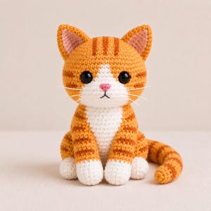 Op de afbeelding: Een handgemaakte gehaakte kat in oranje en wit. De kat heeft grote zwarte ogen, een roze neus en witte snorharen. Het lichaam is oranje met donkerdere oranje strepen. De oren zijn oranje met roze binnenkant. De kat zit met opgerolde staart.