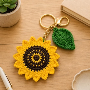 May include: A handmade crochet sunflower keychain with a gold-colored keyring and clasp. The sunflower has yellow petals and a brown center. A green crochet leaf is attached. The keychain is on a wooden surface.