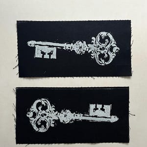 May include: Two black rectangular patches featuring a silver key design. The keys have intricate detailing and are centered on the patches. The patches have a raw, unfinished edge.