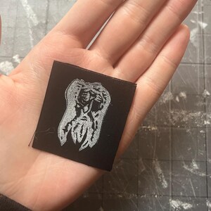 May include: A small, square black patch with a silver embroidered design. The design is an abstract figure or symbol. The patch is held in a hand, showing the scale of the item.