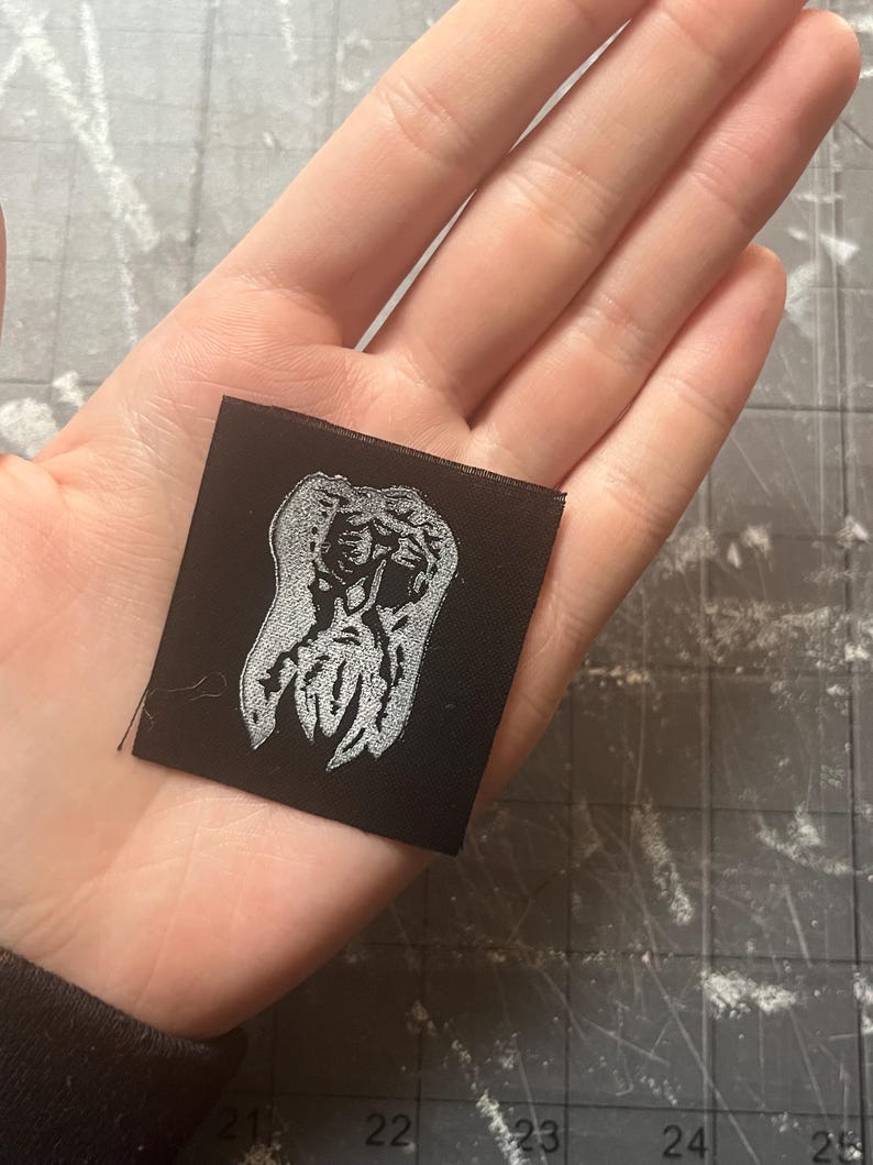 May include: A small, square black patch with a silver embroidered design. The design is an abstract figure or symbol. The patch is held in a hand, showing the scale of the item.