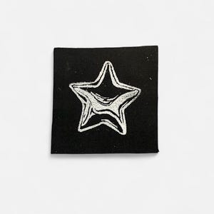 Op de afbeelding: Een zwart vierkant patch met een zilveren sterontwerp. De ster heeft een getextureerd, gemarmerd uiterlijk en is gecentreerd op het vierkant. De patch is waarschijnlijk van stof en kan worden gebruikt voor naai- of knutselprojecten.