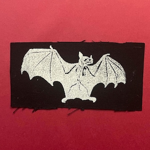 Pu&ograve; includere: Una toppa rettangolare in tessuto nero con una grafica serigrafata bianca di un pipistrello con le ali spiegate. La toppa presenta bordi grezzi leggermente sfilacciati e una trama testurizzata, mostrando un netto contrasto tra lo sfondo scuro e l'inchiostro chiaro.