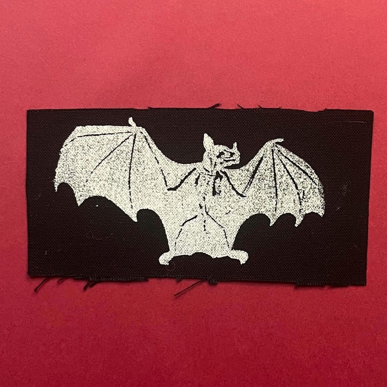 Pu&ograve; includere: Una toppa rettangolare in tessuto nero con una grafica serigrafata bianca di un pipistrello con le ali spiegate. La toppa presenta bordi grezzi leggermente sfilacciati e una trama testurizzata, mostrando un netto contrasto tra lo sfondo scuro e l'inchiostro chiaro.