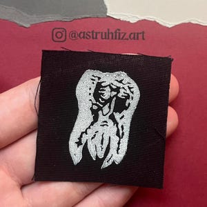 May include: A square black fabric patch featuring a white screen-printed graphic of a stylized tooth. The design has a textured, distressed appearance with visible ink grain. The patch has raw, slightly frayed edges on a dark, woven textile base.