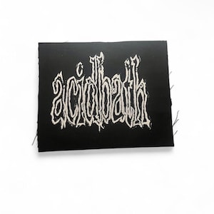 May include: A black square patch featuring the white text "acidbath" in a gothic font. The patch has a rough, textured appearance, possibly fabric. The text is stylized and appears hand-drawn or printed, suggesting a DIY aesthetic.