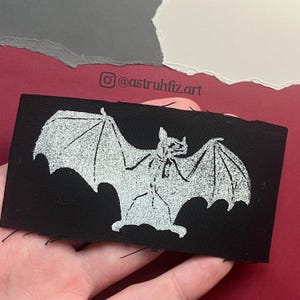 Pu&ograve; includere: Una toppa rettangolare in tessuto nero con una grafica serigrafata bianca di un pipistrello con le ali spiegate. Il design mostra strutture alari dettagliate e un aspetto testurizzato. I bordi della toppa sono grezzi e leggermente sfilacciati.