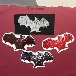 Pu&ograve; includere: Quattro articoli a tema pipistrello con disegni scheletrici. Una toppa rettangolare nera con un pipistrello argentato e tre adesivi fustellati: uno rosso scuro, uno rosso brillante glitterato e uno nero con bianco. Il testo riporta @astruhfiz.art.