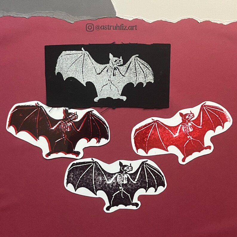 Pu&ograve; includere: Quattro articoli a tema pipistrello con disegni scheletrici. Una toppa rettangolare nera con un pipistrello argentato e tre adesivi fustellati: uno rosso scuro, uno rosso brillante glitterato e uno nero con bianco. Il testo riporta @astruhfiz.art.