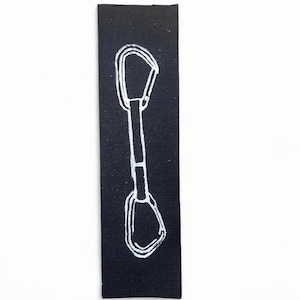 May include: A black rectangular fabric patch with a white outline illustration of a climbing carabiner. The carabiner design is centrally placed on the patch, with a minimalist style. The patch is likely for outdoor enthusiasts.
