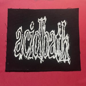 Può includere: Una toppa rettangolare in tessuto nero con la parola acidbath stampata in un carattere bianco stilizzato, frastagliato e dall'aspetto usurato. Le lettere hanno un aspetto disegnato a mano con linee irregolari e bordi grezzi sul tessuto.