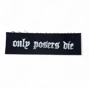 Puede incluir: Un parche rectangular negro con la frase "only posers die" en letras góticas plateadas. El parche es probablemente de tela y está sobre un fondo blanco.