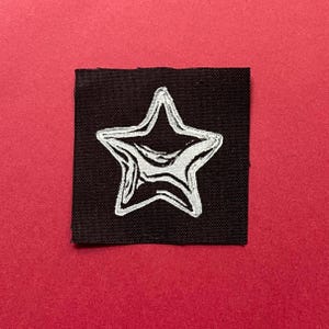 May include: A square black fabric patch featuring a white embroidered star design in the center. The star has a thick, textured outline with internal line details that create a stylized, graphic appearance against the solid black background.