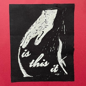 May include: A rectangular black fabric patch featuring a white screen-printed graphic of a hand reaching downward. The design includes the text "is this it" in a stylized white font positioned below the hand, with a textured, distressed aesthetic.