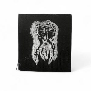 May include: A square, black fabric patch with a white, detailed illustration of an abstract figure. The design has a textured appearance, resembling a creature or mask. The patch is likely for clothing or accessories, and is square in shape.