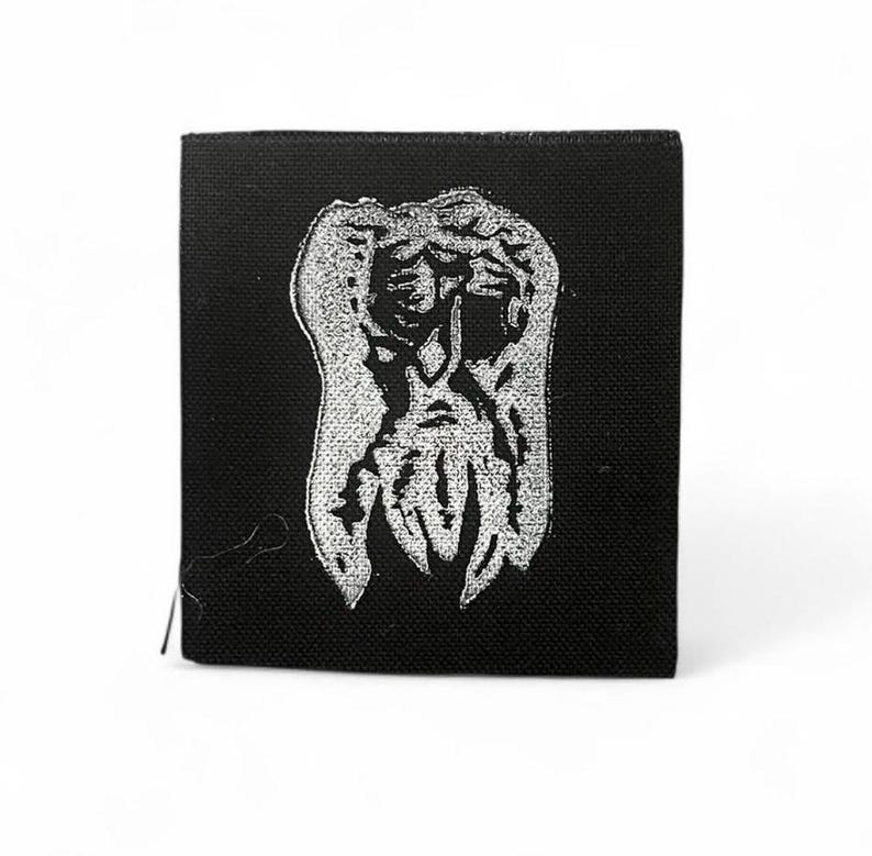 May include: A square, black fabric patch with a white, detailed illustration of an abstract figure. The design has a textured appearance, resembling a creature or mask. The patch is likely for clothing or accessories, and is square in shape.