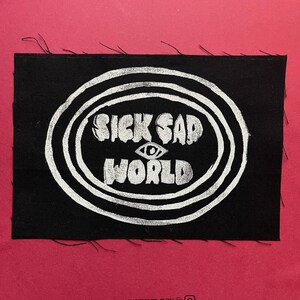 Könnte beinhalten: Ein rechteckiger schwarzer Stoffaufnäher mit ausgefransten Rändern und einem weißen Siebdruckmotiv aus drei konzentrischen Kreisen, die den Text SICK SAD WORLD umgeben, mit einem kleinen Augensymbol zwischen den beiden Textzeilen.