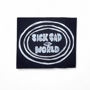 May include: A black square patch with the words "SICK SAD WORLD" in white, stylized text. The text is inside a white oval design with concentric circles. An eye graphic is between "SAD" and "WORLD".