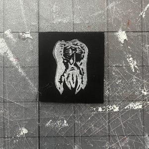 May include: A black square patch with a white, stylised illustration of a face with a beard. The design has a textured appearance. The patch is set against a grey surface with grid lines.