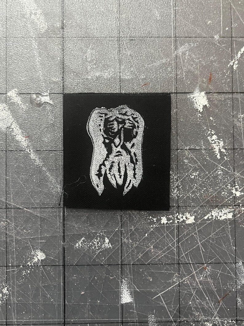May include: A black square patch with a white, stylised illustration of a face with a beard. The design has a textured appearance. The patch is set against a grey surface with grid lines.