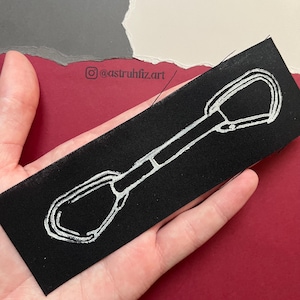 Pode incluir: Um emblema retangular de tecido preto com um desenho de linha branca de uma costura de escalada, que consiste em dois mosquetões conectados por uma fita curta. O design está centralizado no tecido e o texto @astruhfiz.art está no fundo.