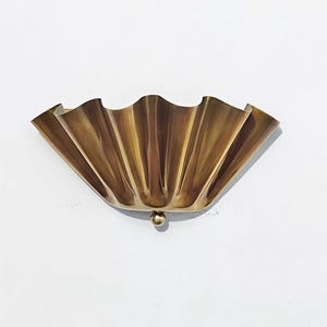 Könnte beinhalten: Eine messingfarbene Wandleuchte mit einem fächerförmigen, gewellten Design. Die Leuchte hat ein kleines, abgerundetes Detail an der Unterseite. Das Metall hat einen warmen, goldenen Farbton, und das Stück ist an einer weißen Wand befestigt.