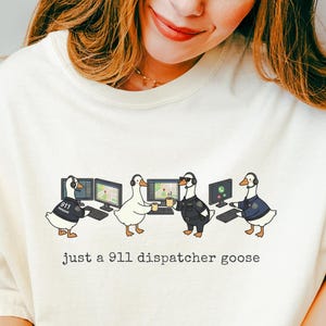Puede incluir: Camiseta color marfil con un diseño de cuatro gansos trabajando en monitores de ordenador. El texto "Just a 911 dispatcher goose" está impreso debajo. La camiseta es de corte holgado.