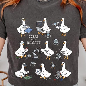 May include: A charcoal gray short-sleeve t-shirt featuring a graphic print of white geese engaged in various technological tasks, including robotics and engineering, with the text "IDEAS into REALITY" printed in the center of the design.