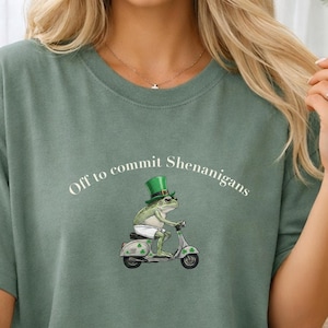 May include: Light green t-shirt featuring the phrase "Off to commit Shenanigans" and a frog on a scooter. The frog wears a green hat, and the scooter has shamrock details. The words "Light Green" are in the upper left corner.