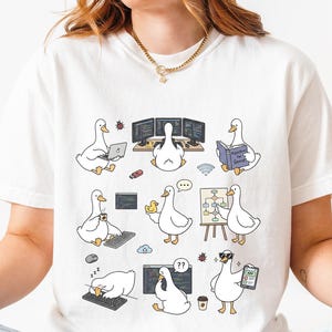 May include: A white short-sleeve t-shirt featuring a graphic print of various cartoon ducks engaged in computer programming and office tasks, including typing on keyboards, reading books, using laptops, and presenting on a whiteboard.