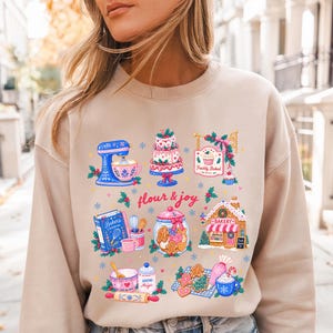 May include: A sand-colored crewneck sweatshirt featuring a colorful graphic print of baking-themed illustrations including a mixer, a tiered cake, a gingerbread house, and the text "flour & joy" surrounded by small blue and red snowflake accents.