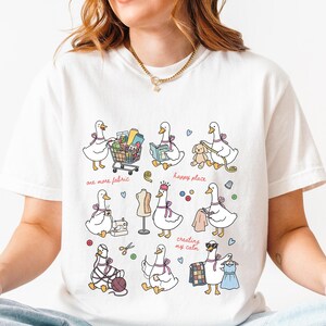 Puede incluir: Una camiseta blanca de manga corta con un estampado gráfico de varios gansos blancos realizando diversas actividades manuales, como costura, tejido y compras. El diseño incluye pequeños iconos coloridos y el texto one more fabric, happy place y creating my calm.