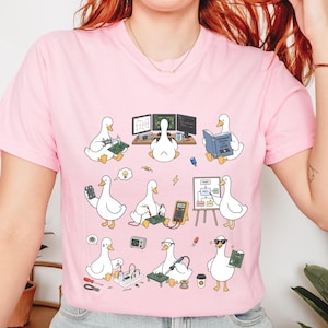 Può includere: Una t-shirt rosa chiaro con una stampa grafica di anatre bianche impegnate in varie attività di ingegneria ed elettronica, tra cui saldatura, uso di un multimetro, lettura di manuali tecnici e lavoro con circuiti stampati e computer.