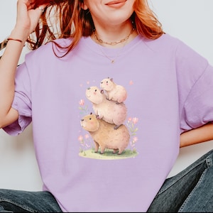 May include: A light purple t-shirt featuring a whimsical illustration of three capybaras stacked on top of each other. The bottom capybara is tan, the middle one is pink, and the top one is light pink. Pink flowers and green grass surround the capybaras.