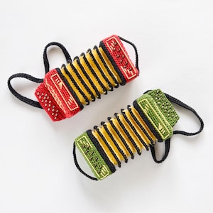 May include: Two miniature crochet accordions, one red and one green, each featuring yellow bellows, black trim, and small metallic beads representing buttons. Both instruments include black crocheted loops on the sides for handling.
