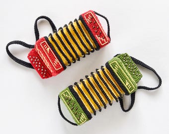 May include: Two miniature crochet accordions, one red and one green, each featuring yellow bellows, black trim, and small metallic beads representing buttons. Both instruments include black crocheted loops on the sides for handling.