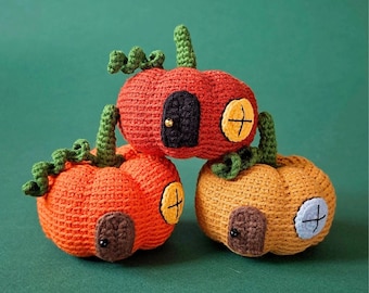 May include: Three crocheted pumpkin-shaped houses in orange, red, and yellow. Each pumpkin features a small dark door, a circular window with a cross-stitch pattern, and a green crocheted stem with curly vines. The textures are soft and yarn-based.
