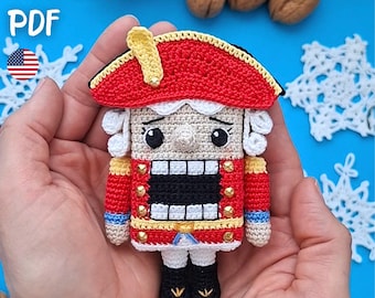 May include: A crochet nutcracker ornament featuring a red hat with a yellow plume, a beige face, and a red jacket with gold accents. The figure has black boots, white hair, and a black rectangular mouth. The text PATTERN PDF is visible.