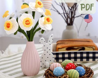 May include: A pink vase holds a bouquet of white and yellow crochet daffodils. A white ceramic bunny with black polka dots and a nest of colorful crochet eggs are on a striped table runner. The image includes text that says "PATTERN PDF".