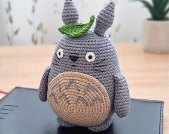 May include: A gray crocheted plush toy shaped like a round creature with two pointed ears, a green leaf on its head, white eyes with black pupils, and a tan belly featuring dark gray chevron patterns. The toy has small arms and feet.