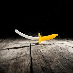 Könnte beinhalten: Ein skurriles, bananenförmiges Schwert mit gelber Klinge und weißem Griff liegt auf einer verwitterten Holzoberfläche. Das verspielte Design hat einen gelben Knauf und eine gebogene Klinge, was ein einzigartiges Stück ergibt.