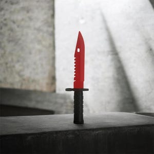 Puede incluir: Un cuchillo rojo y negro con un borde dentado y una punta. El cuchillo tiene un mango negro y una hoja roja. El cuchillo está de pie sobre una superficie oscura.