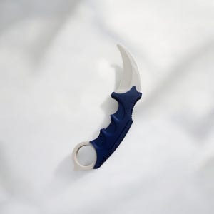 May include: A curved karambit knife. It has a white blade and a dark blue handle with finger grooves and a ring at the base. The knife is set against a white background.