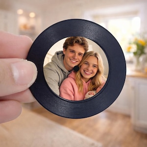 May include: A dark blue, circular photo frame with a textured outer edge. The frame contains a colour photograph of a smiling couple. The background is blurred, suggesting an indoor setting. The frame is held by a hand.