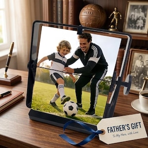 May include: A black rotating picture frame displaying a photograph of an adult and a child playing soccer on a grass field. A white gift tag with a blue ribbon is attached to the frame, featuring the text "FATHER'S GIFT To My Hero, with Love."