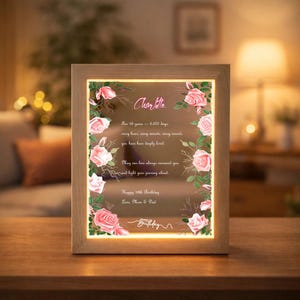 Peut inclure: Un cadre en bois lumineux rectangulaire avec un panneau en verre orné de roses roses et de feuilles vertes sur les bords. Le texte central indique : Charlotte, For 18 years - 6,570 days, every hour, every minute, every second, you have been deeply loved.